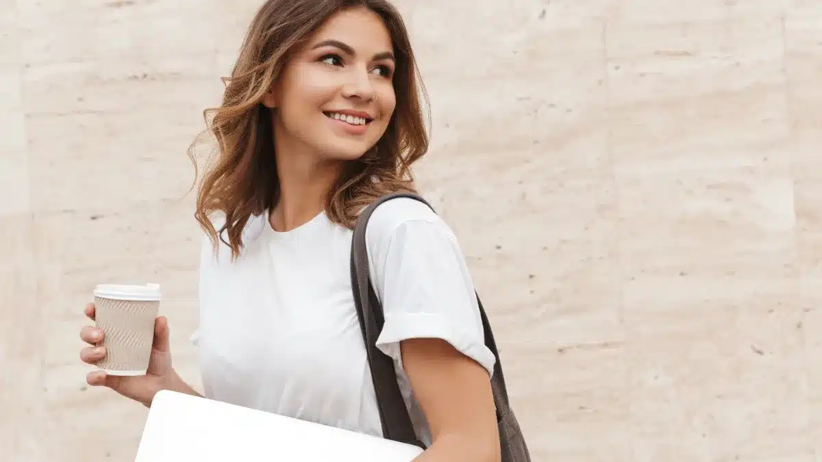 Tânără europeană mergând încrezătoare cu laptop și cafea în mâini