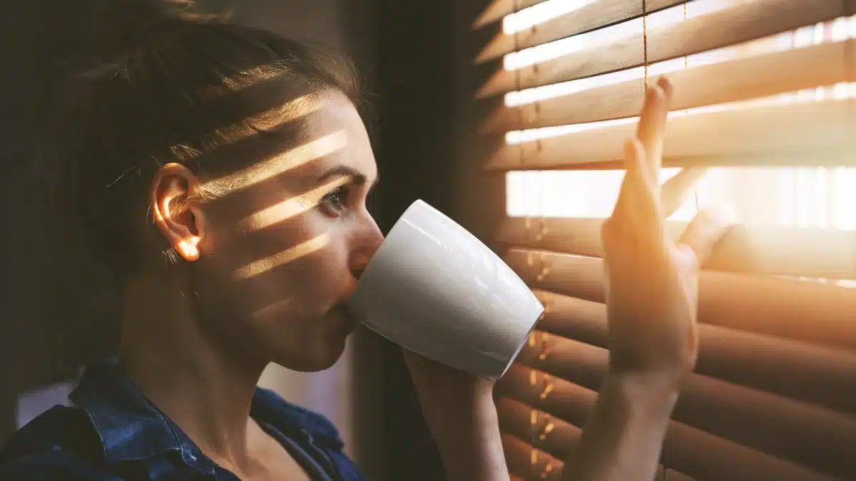Femeie care privește pe fereastră în lumină naturală, cu o cană de cafea în mână