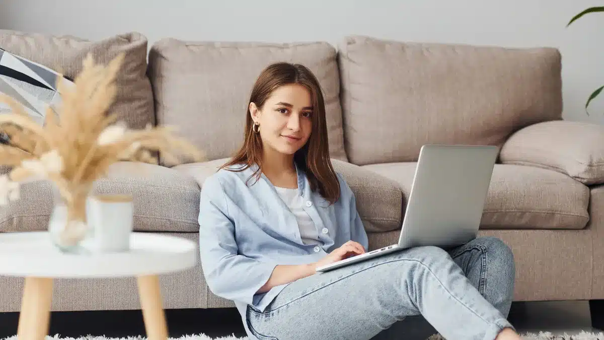 Tânără care lucrează de acasă, cu laptopul, într-un living luminos — comparația între videochat de acasă și din studio